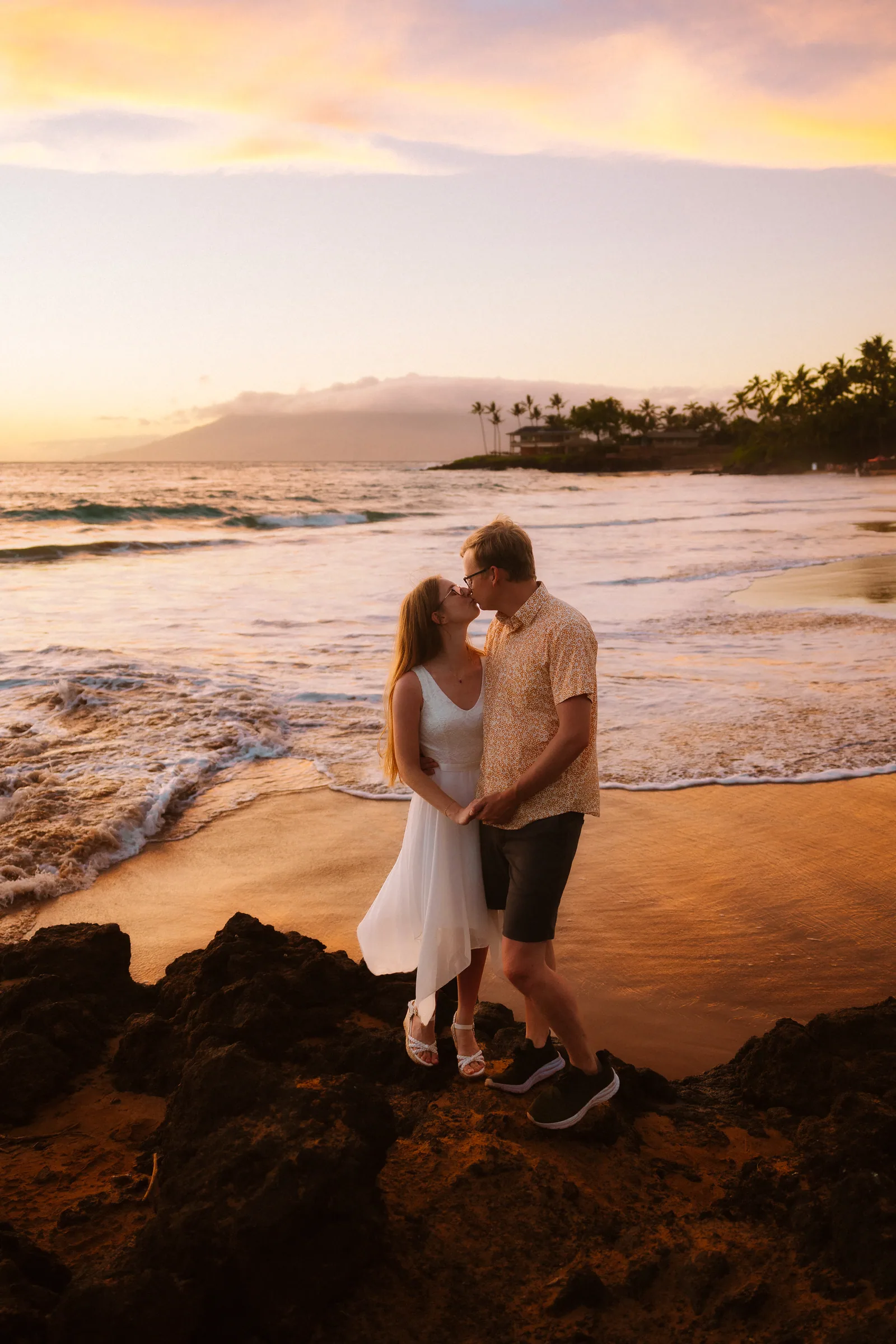 A couple embracing on the sand with rocks and a pastel sky behind them