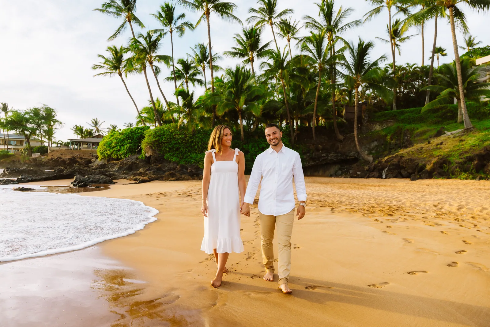 A couple in white walking the shoreline beneath a stand of coconut palms