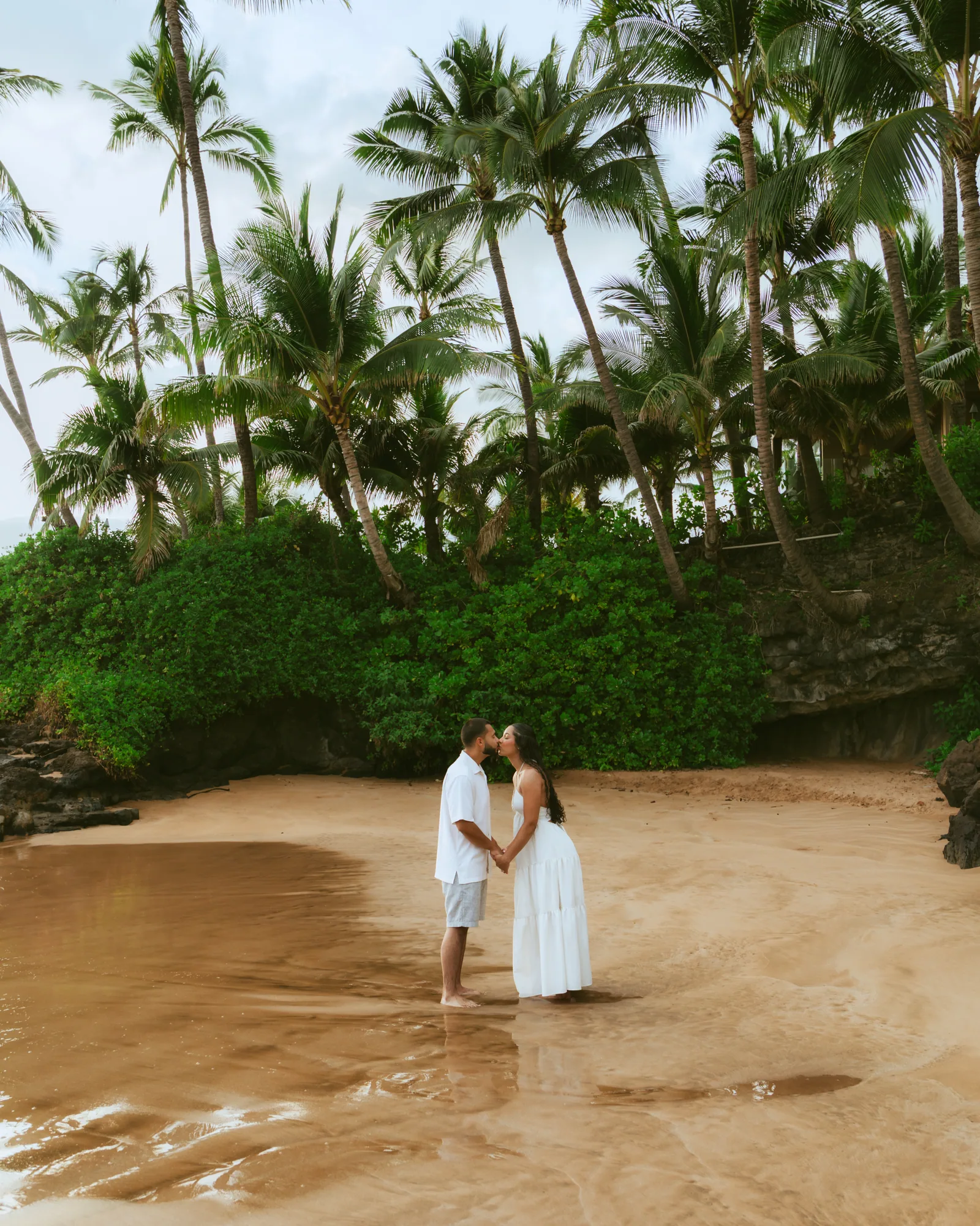 A couple in white kissing at the water's edge, photographed from a distance